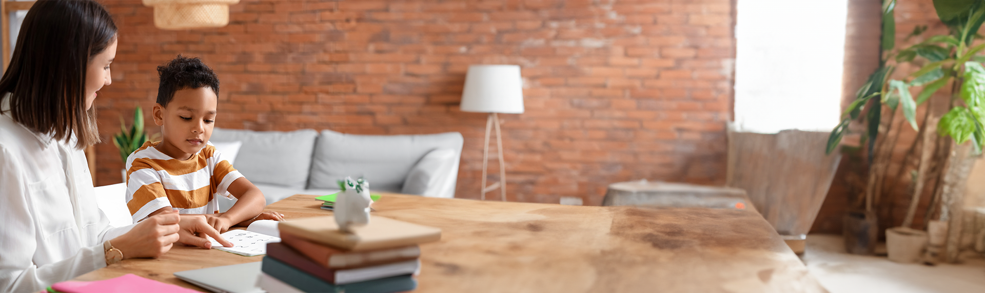 A woman, who works as a Criminal Defense Lawyer, helps a child with homework at a wooden table. Books and a toy are scattered about. The room exudes coziness with brick walls, a soft gray sofa, a floor lamp, and a large plant in the background.