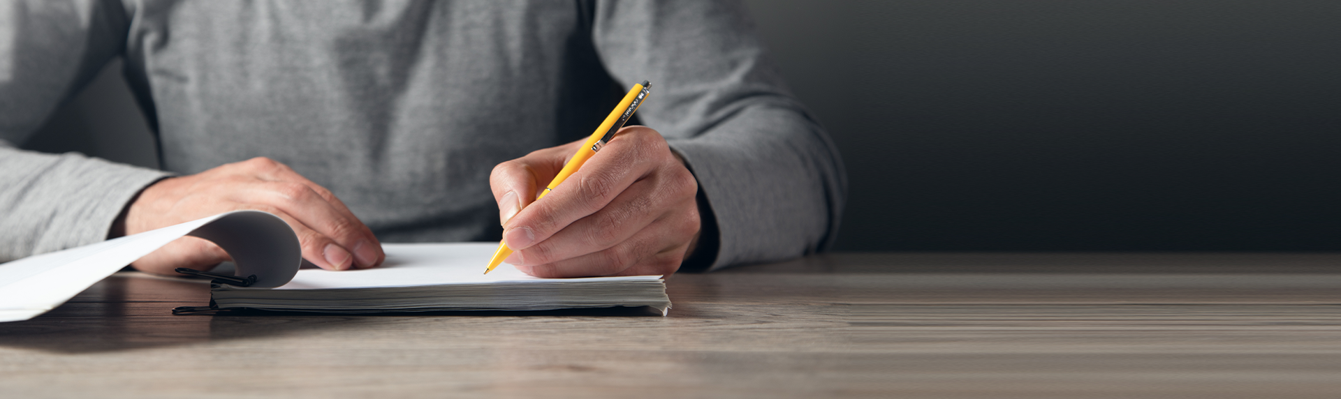 A person in a gray shirt, possibly a criminal defense lawyer, writes with a yellow pencil on a notepad, resting on a wooden table. The focus is on their hands and the notepad, with the background softly blurred.