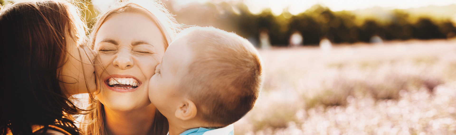 In a sunlit field, a joyful moment unfolds as two children plant kisses on the cheeks of a smiling woman, their mother. The scene encapsulates the warmth and love that mothers' rights attorneys work tirelessly to support. Blurred greenery and warm sunlight form the backdrop of this tender scene.
